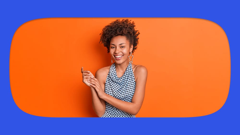 Jovem mulher africana sorridente com cabelo cacheado, vestindo blusa azul de estampa de bolinhas, em fundo de cores vibrantes laranja e azul, mostrando alegria e estilo.