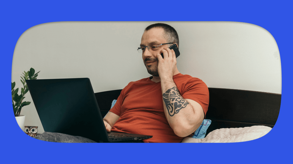 Homem usando celular e escrevendo no laptop, sentado na cama, com uma planta ao lado e fundo azul,consultando o registrato.
