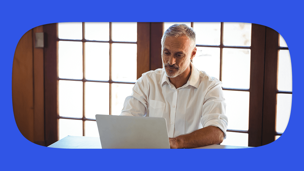 Homem idoso com cabelo grisalho, usando camisa branca, sentado em mesa com notebook consultando tempo de contribuição do INSS, em ambiente bem iluminado, com janelas de vidro ao fundo.