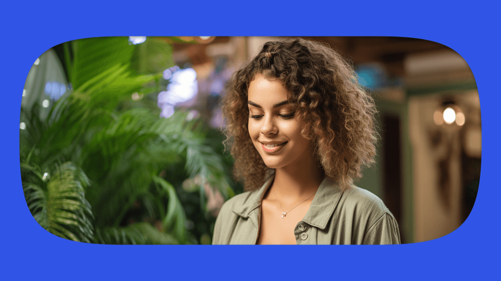 Jovem mulher sorridente com cabelo cacheado, usando jaqueta leve, em ambiente com plantas verdes ao fundo.