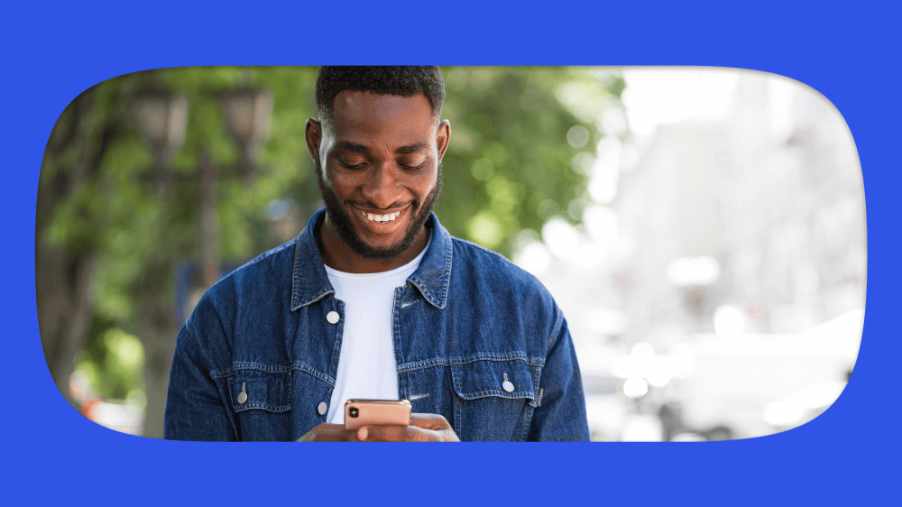 Homem sorridente usando smartphone para calcular o décimo terceiro na calculadora grátis do Pagou Fácil, com fundo de árvores e céu claro.