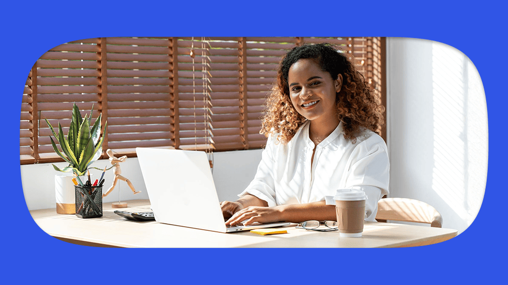 Jovem mulher de cabelos cacheados e sorriso no rosto consultando dataprev pelo cpf em um laptop em um escritório bem iluminado com plantas e decoração moderna.