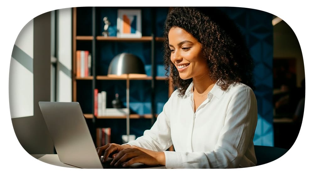 Mulher sorridente usando laptop em um escritório moderno, com decoração de livros e objetos de design industrial ao fundo.