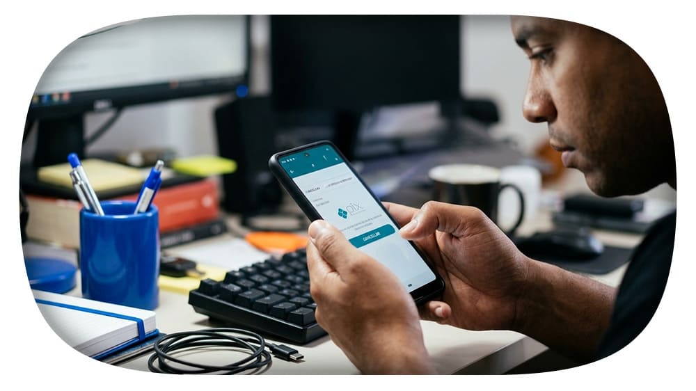 Homem usando um smartphone em um escritório com teclado, canetas e outros materiais de escritório ao redor, voltado para a tecnologia móvel.