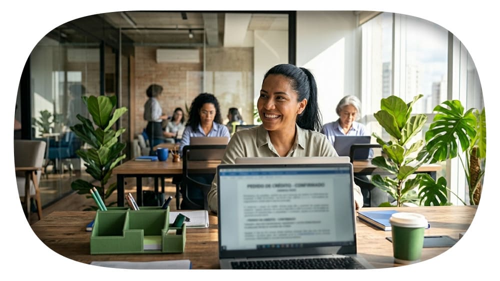 Mulher sorridente trabalhando em um escritório moderno com mesas, notebook, xícara de café e plantas ao lado, com outras pessoas desfocadas ao fundo.