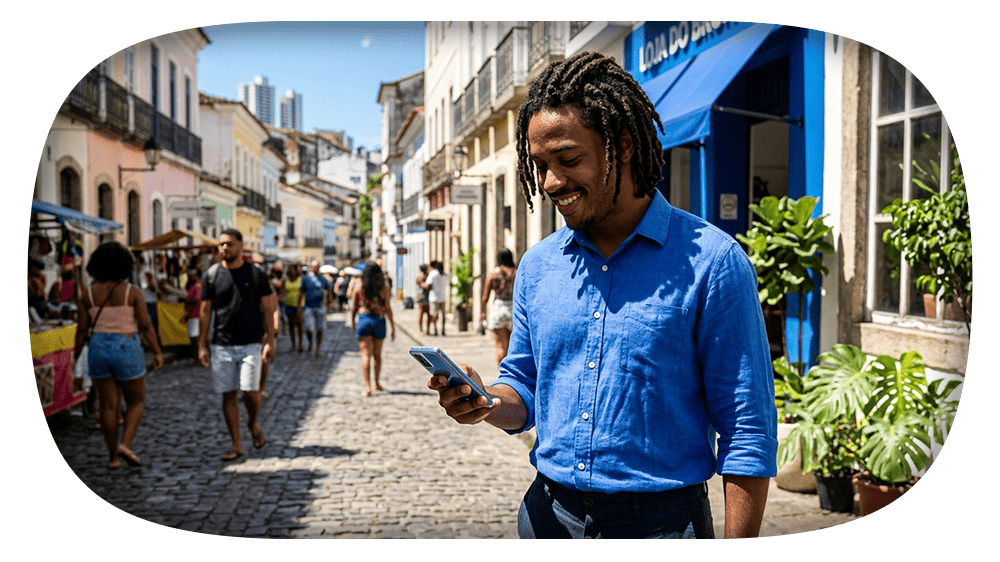 Homem com camisa azul em rua comercial de paralelepípedos, sorrindo e usando o celular enquanto pedestres caminham ao fundo.