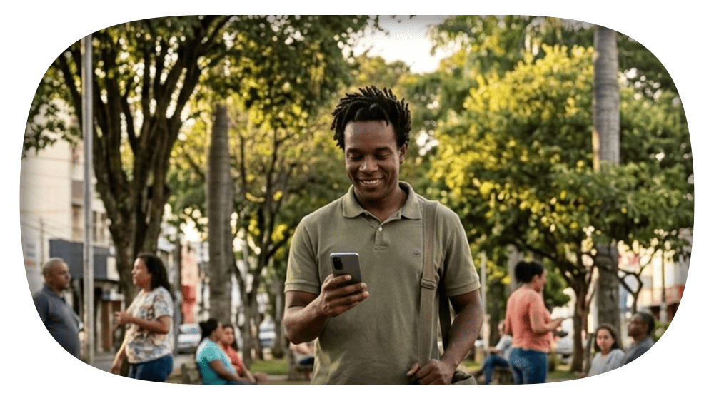 Homem jovem caminhando na rua arborizada ao entardecer, sorrindo e usando o celular enquanto pessoas passam ao fundo.