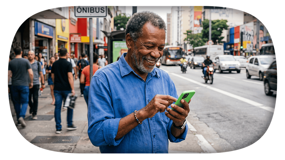 Homem mais velho sorrindo enquanto usa o celular em uma avenida movimentada da cidade, com pedestres, ônibus e carros ao fundo.