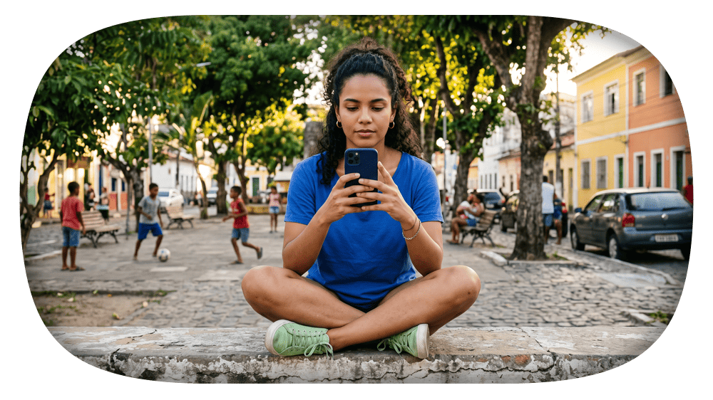 Mulher sentada com pernas cruzadas em uma calçada de pedras, segurando o celular e fotografando-se em um passeio ao ar livre, com pessoas e prédios ao fundo.