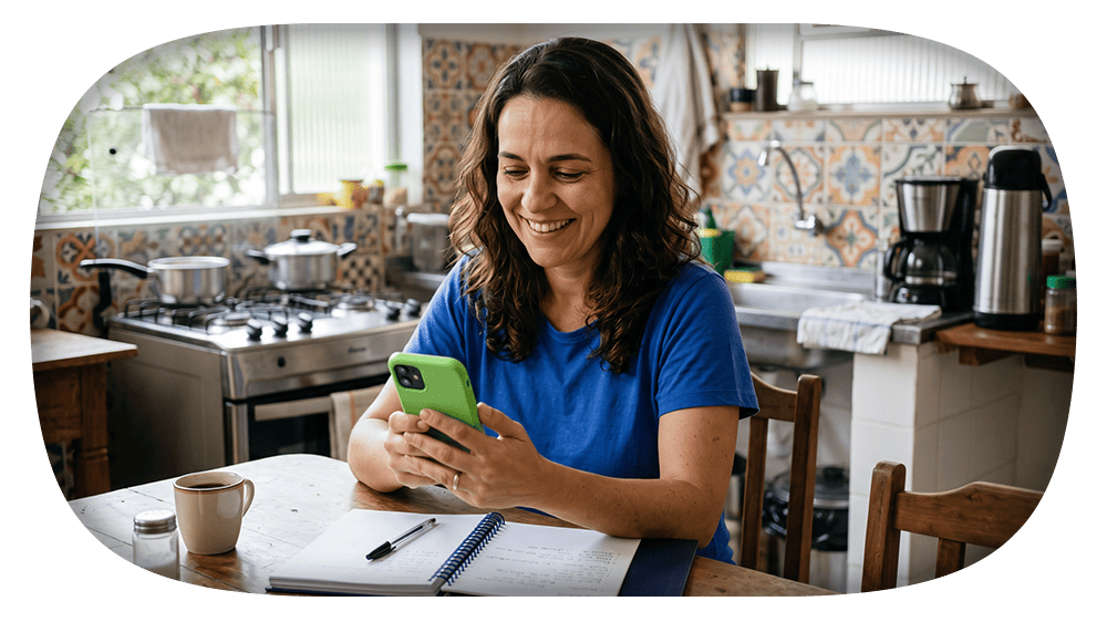 Mulher sorrindo na cozinha usando celular verde e fazendo anotações em um caderno sobre a mesa, com cafeteira ao fundo e fogão ao lado, em ambiente iluminado