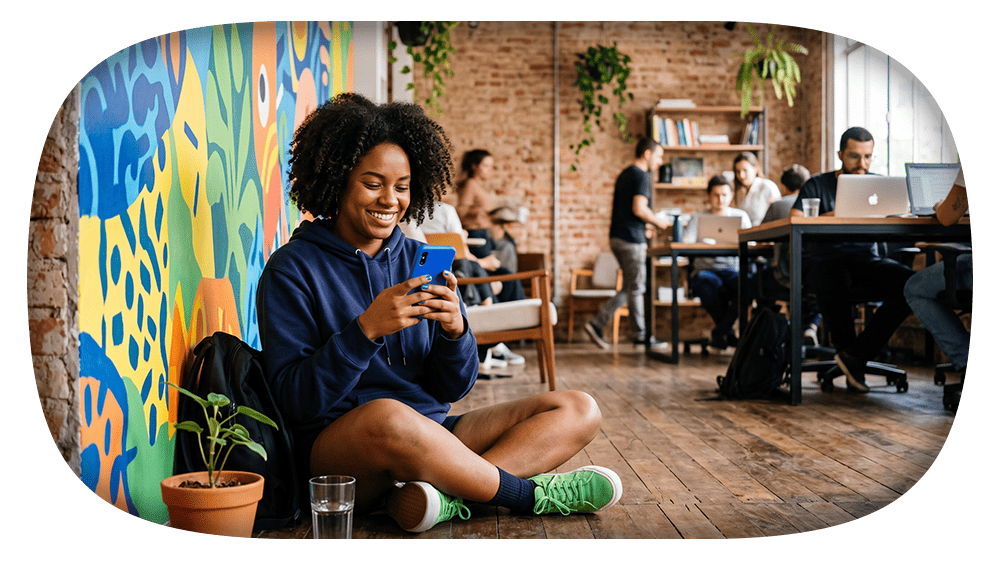 Jovem mulher sentada no chão em um ambiente de coworking, sorrindo enquanto usa o celular e segura um cartão azul, ao lado de uma plantinha em vaso e pessoas trabalhando ao fundo.
